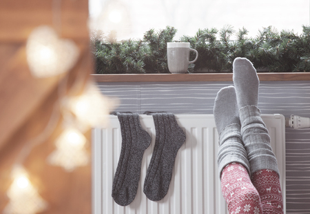 49400436 - woman warming up with feet on heater winter woolen socks drying on a heater, christmas lights, decorations and hot drink
